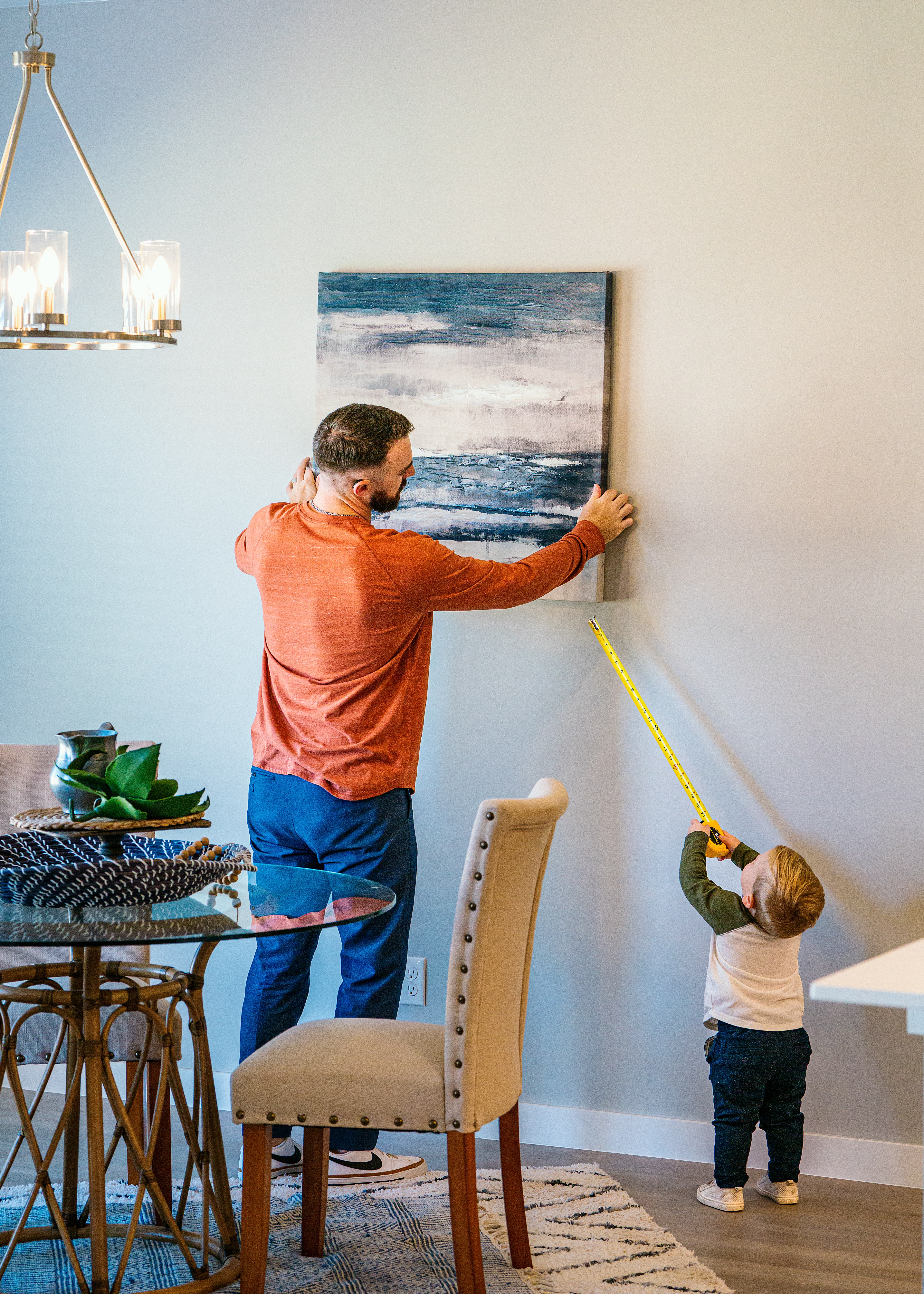 Small child holding a measuring tape up to a painting as his father hangs it on the wall